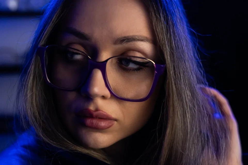 Junge Frau mit kräftig violetter Kunststoffbrille im Profil, blaues Gegenlicht auf dem dunklen Haar.