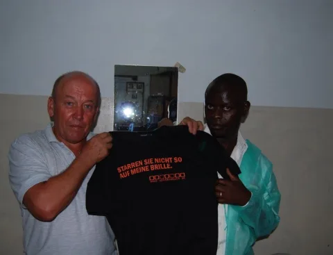 Zwei Männer halten gemeinsam ein schwarzes T-Shirt mit der Aufschrift „Stadion sie nicht so auf meine Brille“ in die Kamera