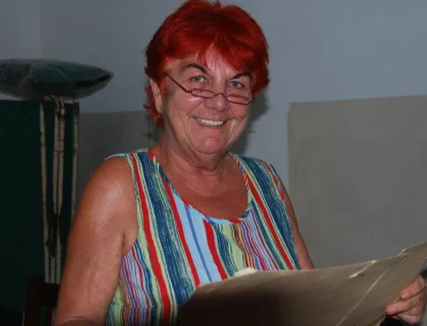 Eine Frau mit roten Haaren und Brille lächelt in die Kamera, in einem Raum mit Untersuchungsliege