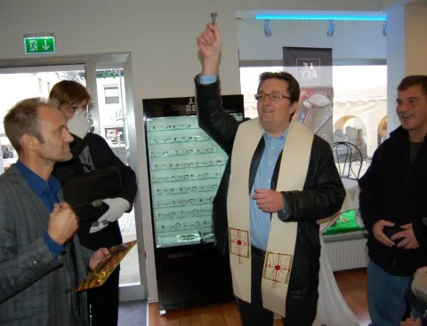 Ein Priester mit Stola segnet den Verkaufsraum, daneben ein Ministrant mit Weihrauchfass vor einer Brillenvitrine.