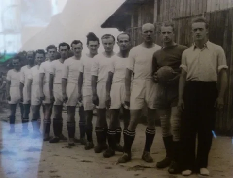 Historische Schwarzweißaufnahme einer Fußballmannschaft, aufgereiht vor einer Holzwand.