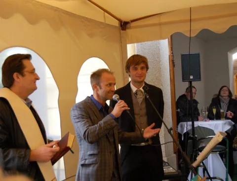 Ansprache im Festzelt: Ein junger Mann spricht ins Mikrofon, daneben der Priester.