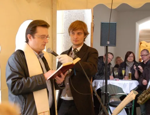 Der Priester liest aus einem Buch, daneben ein junger Mann mit Mikrofon.