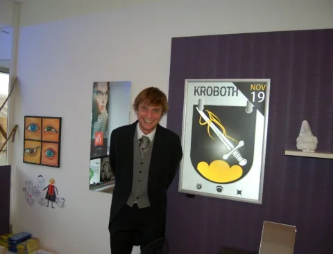Ein junger Mann steht neben dem gerahmten Kroboth-Wappen an der violetten Wand.