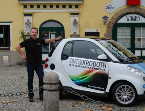 Ein Mitarbeiter zeigt auf den beschrifteten Firmen-Smart vor dem Geschäft.