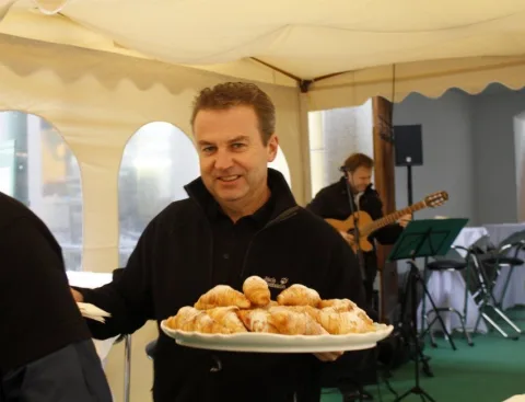 Ein Mitarbeiter bringt eine Platte mit Gebäck, im Hintergrund die Musiker.