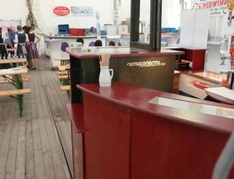 Blick über die geschwungene rote Bartheke in ein Festzelt mit Bierbänken, im Hintergrund Gäste