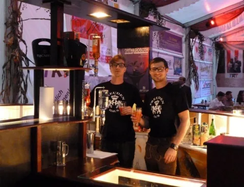 Zwei Personen in schwarzen T-Shirts stehen hinter der beleuchteten Bar zwischen Gläsern und Flaschen