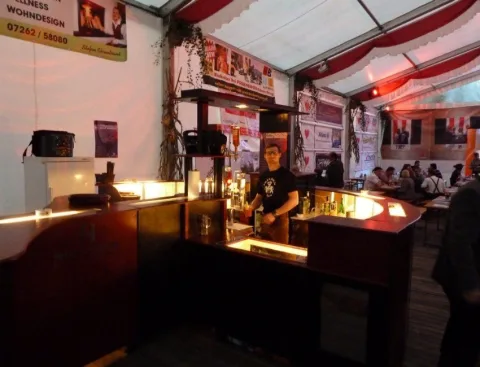 Die beleuchtete Bar im Festzelt von der Seite, dahinter eine Person, ringsum Werbebanner