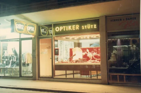 Eine alte Aufnahme eines Geschäfts bei Nacht: beleuchtete Schaufenster unter dem Schriftzug „Optiker Stütz"