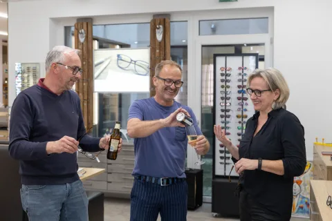 Drei Personen stoßen im Verkaufsraum miteinander an, einer hält eine Flasche