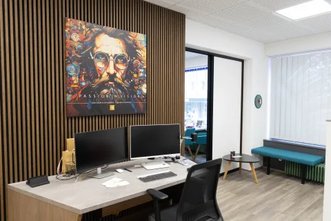 Ein Arbeitsplatz mit zwei Bildschirmen vor einer Holzlamellenwand, darüber ein großes Pop-Art-Bild mit Brillenmotiv, rechts eine türkise Sitzbank am Fenster