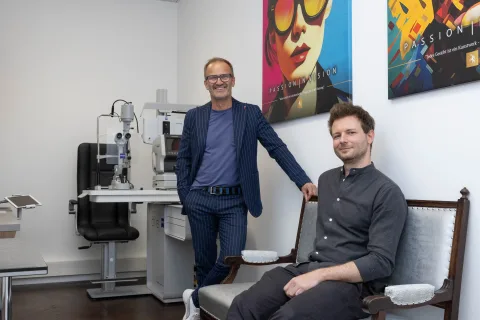 Zwei Augenoptiker im Untersuchungsraum: einer steht an der Spaltlampe, der andere sitzt im Behandlungsstuhl