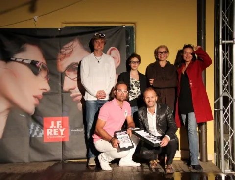 Eine Gruppe vor einer J.F.-Rey-Wand, ein Mann kniet mit einem Brillentablett