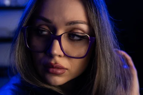 Junge Frau mit kräftig violetter Kunststoffbrille im Profil, blaues Gegenlicht auf dem dunklen Haar.