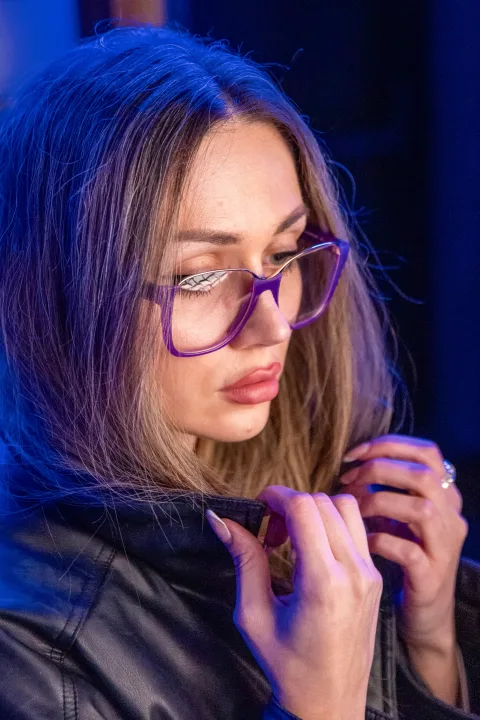 Frau mit violetter Brille in blauem Licht, den Blick gesenkt, die Hände am Kragen.