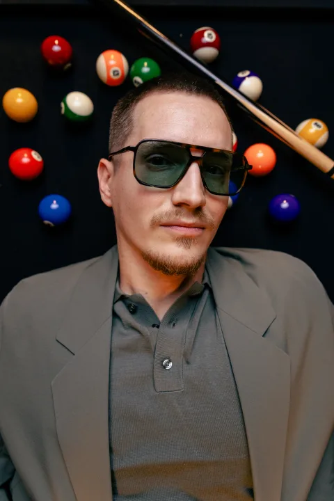 Mann mit dunkler Sonnenbrille vor einer Billardwand, hinter ihm die bunten Kugeln.