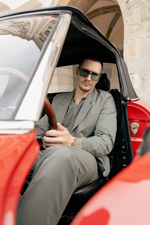 Mann mit Sonnenbrille sitzt in der offenen Tür eines roten Sportwagens.