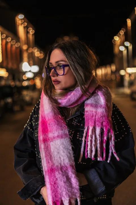 Frau mit Brille und rosa Schal in einer nächtlichen Gasse, Lichterketten im Hintergrund.