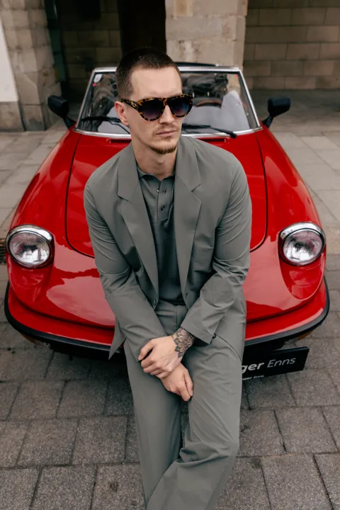 Mann im grauen Anzug mit Sonnenbrille sitzt auf der Motorhaube eines roten Sportwagens.
