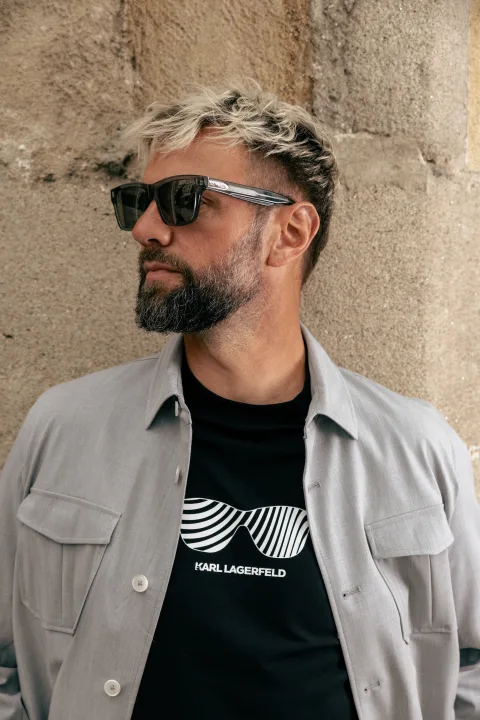 Mann mit heller Jacke und Sonnenbrille vor einer alten Steinmauer.
