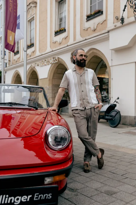 Mann mit Sonnenbrille geht an einem roten Oldtimer vorbei, dahinter bunte Altstadthäuser.