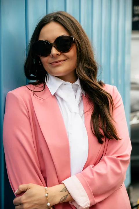 Frau im rosa Blazer mit großer Sonnenbrille vor einer hellblauen Wand.