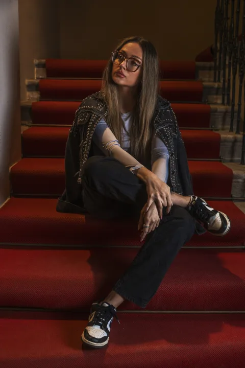 Frau mit Brille sitzt auf der Treppe mit rotem Läufer, das Licht fällt von der Seite.