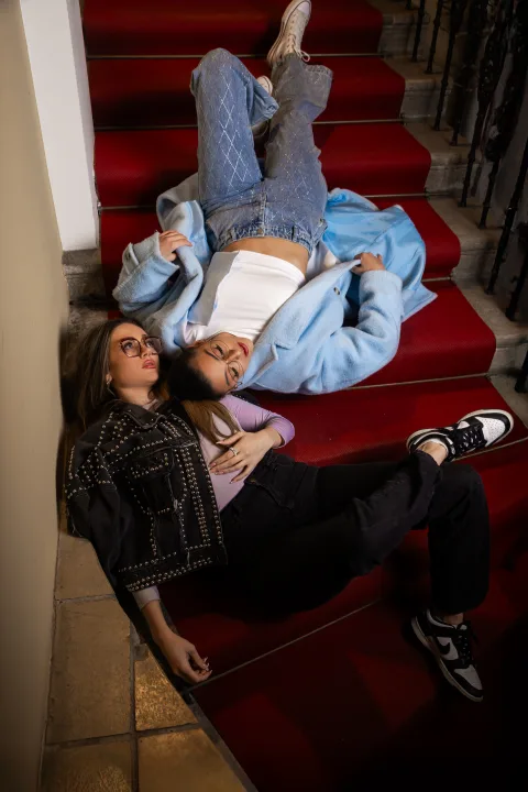 Zwei junge Frauen liegen lachend auf der roten Treppe, eine hält ein Buch.