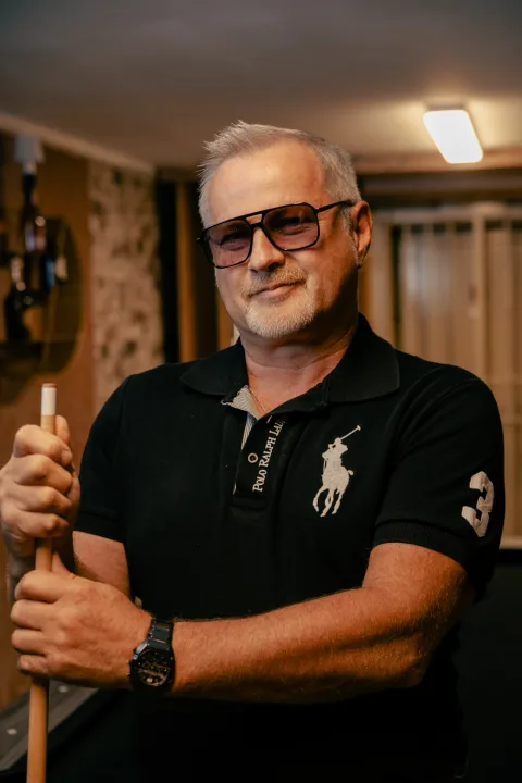 Mann mit grauem Haar, Bart und Brille im schwarzen Polohemd, warmes Licht.