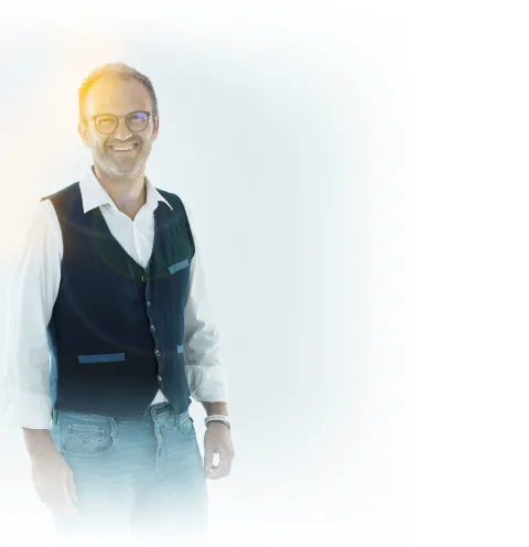 Ein Mitarbeiter mit Brille und grauem Haar, in Hemd und dunkler Weste, vor weißem Hintergrund