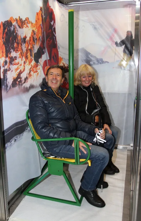 Ein Paar sitzt im Sessellift vor der Skifahrer-Kulisse.