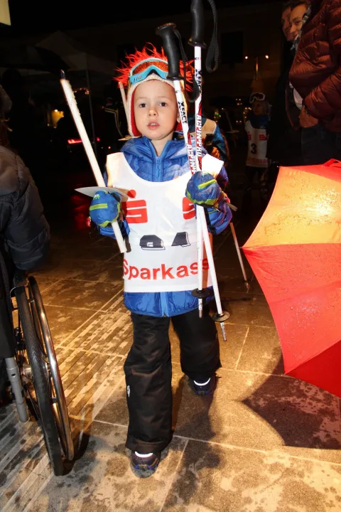 Ein Kind in Skirennanzug mit Startnummer und Skistöcken.