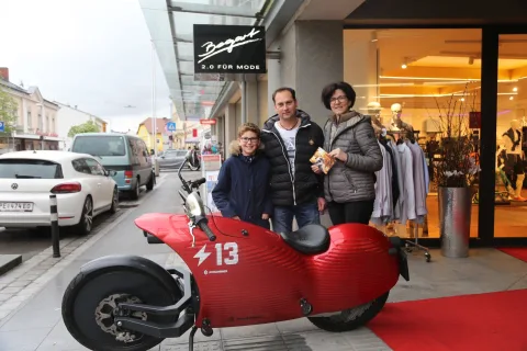 Ein rotes Elektro-Motorrad steht auf einem Teppich vor einem Modegeschäft, davor drei Personen
