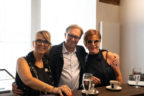 Zwei Frauen und ein Mann stehen eng beieinander an einem Tisch mit Gläsern