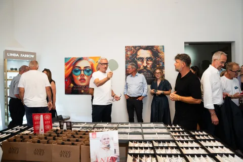 Gäste stehen im Geschäft um einen langen Tisch voller Brillen, an den Wänden Pop-Art-Bilder