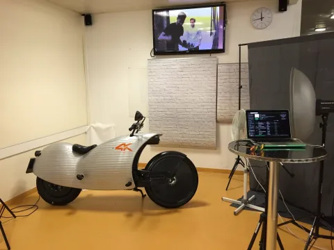 Ein weißes Elektro-Motorrad steht auf gelbem Boden neben einem Stehtisch mit Laptop