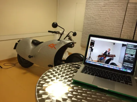 Ein weißes Elektro-Motorrad neben einem Stehtisch, auf dem ein Laptop ein Video zeigt