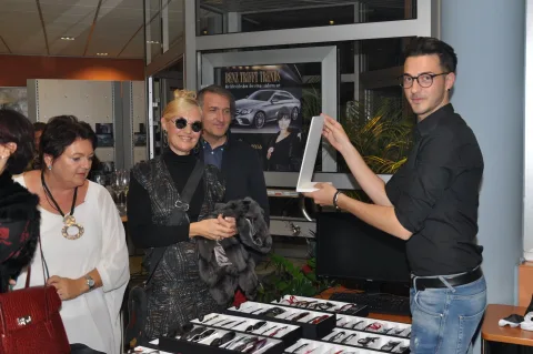 Ein Mitarbeiter zeigt zwei Besucherinnen eine Brille, im Hintergrund ein Autohaus-Schauraum