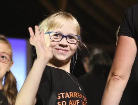 Ein Mädchen mit Brille winkt in die Kamera