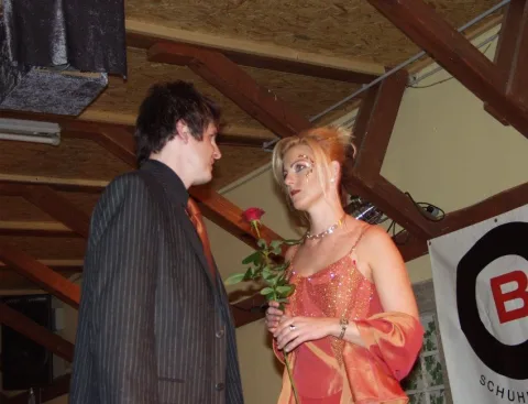 Ein Model im Anzug überreicht einer Frau im orangefarbenen Abendkleid eine Rose