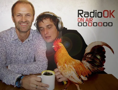 Zwei Männer vor einer Aufstellwand von Radio OK, einer mit Kopfhörern, daneben ein Plakat mit einem Hahn