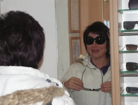 Eine Frau mit dunkler Sonnenbrille probiert vor einer Fassungswand eine zweite Brille