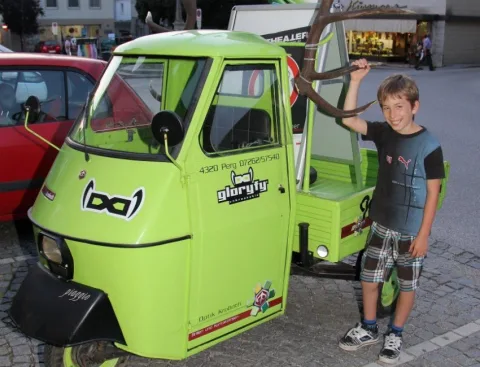 Ein Junge neben einem hellgrünen Dreirad-Kleintransporter auf dem Platz