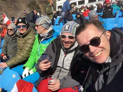Zwei Männer mit Sonnenbrillen im Zuschauerbereich einer Wintersportarena, dicht gedrängt zwischen anderen Fans