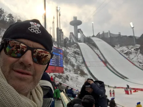 Ein Mann mit verspiegelter Sonnenbrille und Olympia-Mütze vor der Skisprungschanze