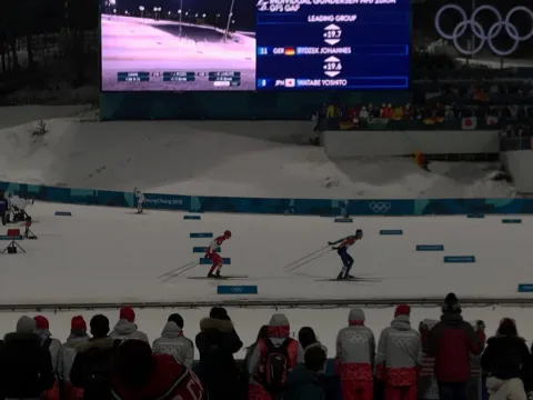 Blick auf die Langlaufstrecke im Stadion, davor Zuschauer, im Hintergrund die Anzeigetafel mit den olympischen Ringen