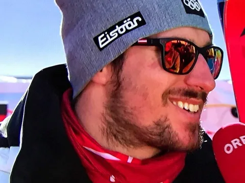 Nahaufnahme eines Sportlers mit Sonnenbrille und Haube, im Hintergrund verschneite Berge