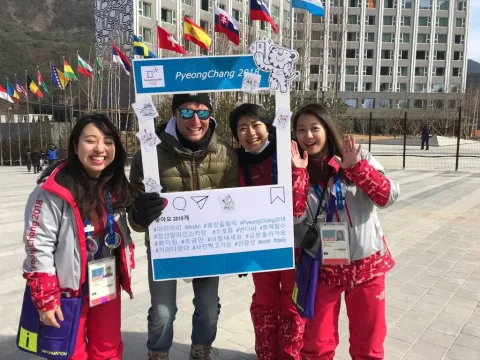 Vier Personen halten einen Fotorahmen mit der Aufschrift PyeongChang in die Kamera, dahinter Fahnenmasten