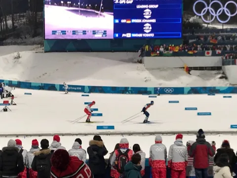 Langläufer auf der Strecke im Stadion, davor die Zuschauerreihen und die Anzeigetafel